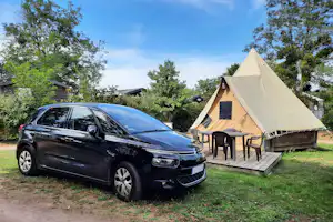 Campingzelt mit Terrasse und Sitzgruppe, daneben ein Auto auf dem Stellplatz im Grünen zwischen Bäumen.