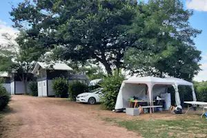 Campingzelt mit Vorzelt und Tisch, daneben ein Auto; im Hintergrund Holzgebäude und große Bäume auf dem Stellplatz.