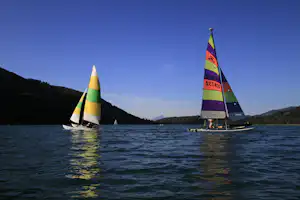 Zwei Segelboote auf dem See, bunte Segel im Vordergrund, bewaldete Ufer und blauer Himmel im Hintergrund.