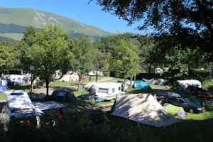 Campingplatz mit Wohnwagen und Zelten zwischen Bäumen, Parkplatz und Weg, im Hintergrund grüne Berge.
