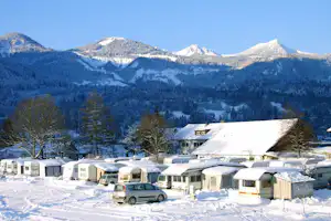 Campingplatz im Winter: Wohnwagen und Mobilheime auf verschneitem Gelände, im Hintergrund Berge und ein großes Gebäude.