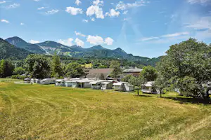 Campingplatz mit Wohnwagen und Zelten auf einer Wiese, im Hintergrund Berge und bewaldete Hänge unter blauem Himmel.
