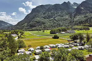 Campingplatz mit Wohnwagen und Zelten inmitten grüner Wiesen, umgeben von Bäumen und hohen Bergen im Tal.