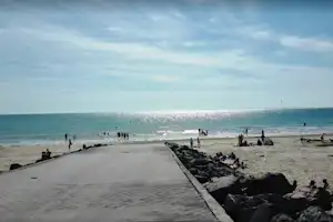 Strand mit Meer und Wellen, dahinter Menschen am Ufer; links eine lange Mauer aus Steinen.