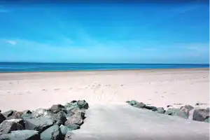 Sandstrand mit Kiesweg zum Meer, Felsbrocken am Rand und Blick auf die Küste unter klarem Himmel.