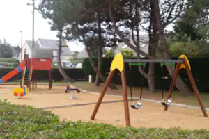 Schaukel und Klettergerüst auf dem Spielplatz am Campingplatz, mit Sandfläche, Bäumen und Wohnhäusern im Hintergrund.