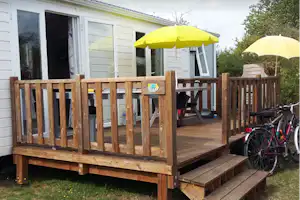 Bungalow mit Holzterrasse, zwei Sonnenschirmen und Fahrrädern am Geländer, vor weißen Wänden und Fenstern.