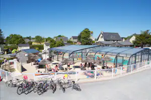 Überdachter Poolbereich mit vielen Fahrrädern davor, Sonnenschirme und Liegen, im Hintergrund Häuser und Bäume.