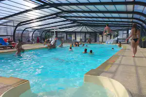 Überdachtes Camping-Schwimmbecken mit blauem Wasser, Liegen und mehreren Badegästen unter einem Glasdach.