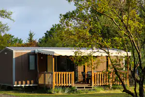Modernes Mobilheim mit Holzveranda und Markise, umgeben von Bäumen und Rasen auf dem Campingplatz.