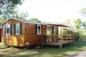 Holz-Mobilheim mit Veranda und Sitzgruppe, umgeben von Rasen und Bäumen im Campingbereich.