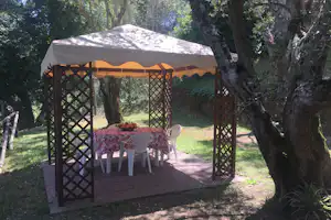 Camping-Pavillon mit Tisch und Stühlen auf einer gepflasterten Terrasse, umgeben von Bäumen und grünem Rasen.