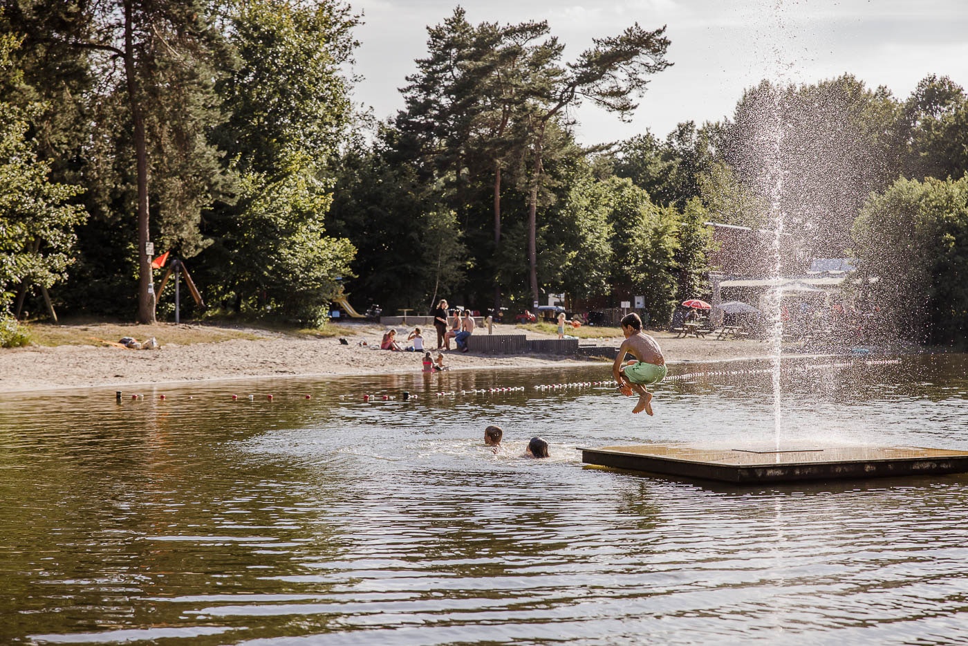 Badesee mit Wasserfontäne und Holzsteg, im Hintergrund Bäume und ein paar Personen am Ufer.