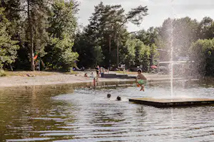 Badesee mit Wasserfontäne und Holzsteg, im Hintergrund Bäume und ein paar Personen am Ufer.