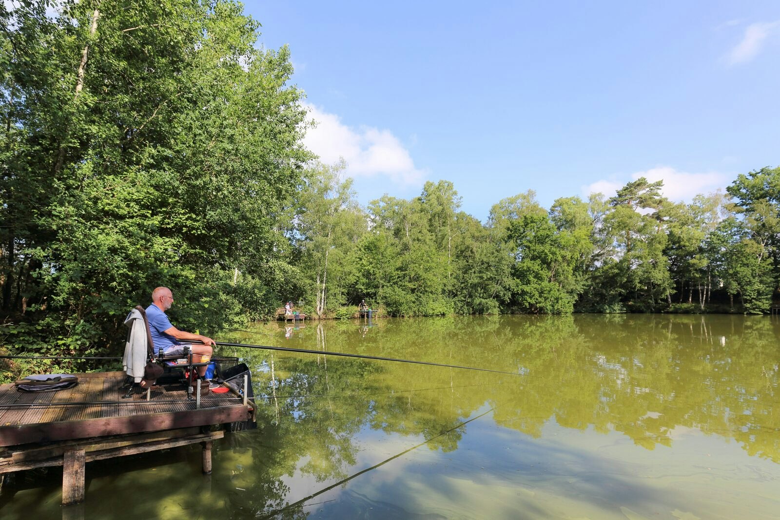 Angler auf einer Holzplattform am See, mit Rutenhalter und Blick auf ruhiges Wasser und dichten Wald.