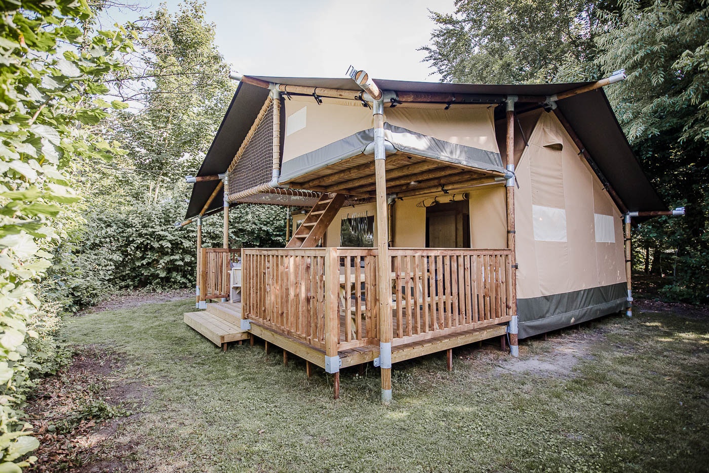 Glamping-Zelt auf Holzpodest mit Veranda, Holzgeländer und Markise, umgeben von Bäumen und Rasenfläche.