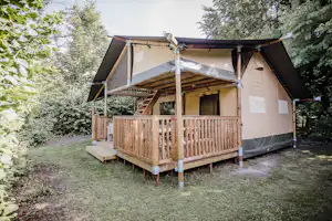 Glamping-Zelt auf Holzpodest mit Veranda, Holzgeländer und Markise, umgeben von Bäumen und Rasenfläche.