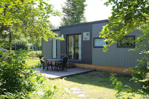 Bungalow mit Holzterrasse und Sitzgruppe vor grauer Fassade, umgeben von Bäumen und grünem Rasen im Campingbereich.