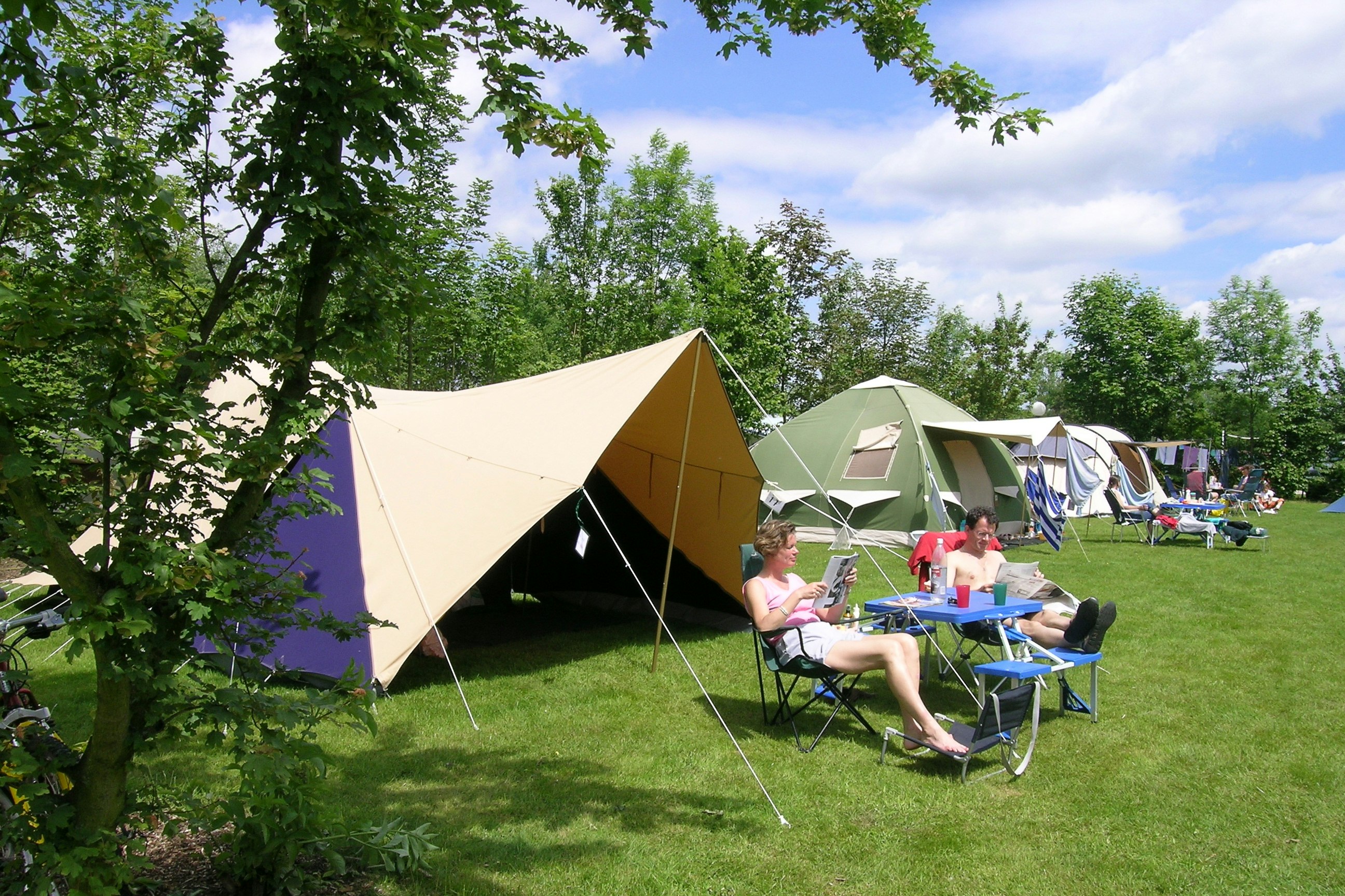 Campingzelte auf grünem Rasen, daneben Campingstühle und Tisch mit Getränken, im Hintergrund weitere Zelte und Bäume.