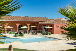 Pool auf der Terrasse mit Sonnenliegen und Sonnenschirmen vor einem roten Campinggebäude, eingerahmt von Palmenblättern.