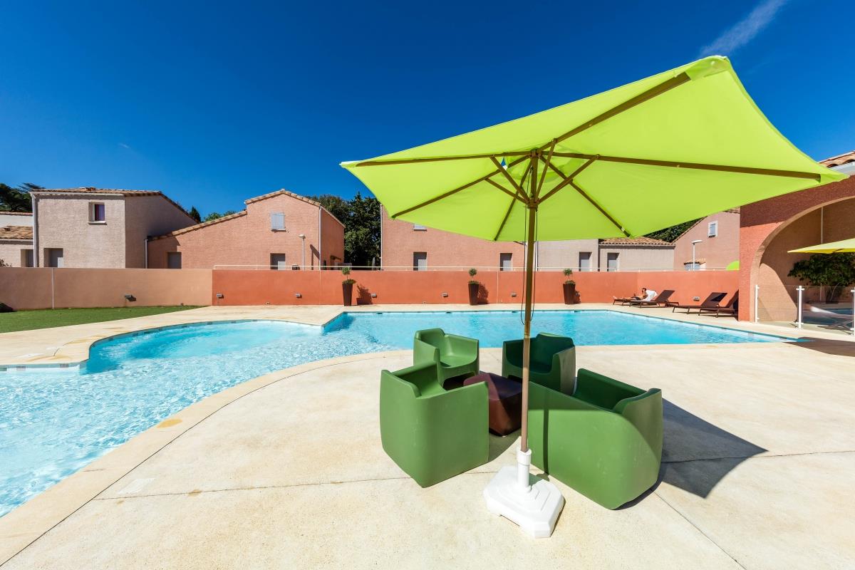 Grüner Sonnenschirm über dem Poolbereich, daneben grüne Liegestühle auf der Terrasse vor den Camping-Gebäuden.