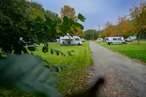 Wohnmobile auf einem Campingplatz entlang eines Schotterwegs, umgeben von Rasen und Bäumen.
