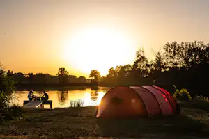 Zelt am Flussufer bei Sonnenuntergang, ruhiges Wasser, Bäume und ein kleiner Steg im Hintergrund.