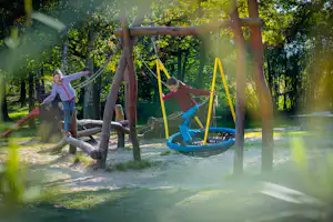 Schaukeln im Campingwald, zwei Kinder auf einer Hängematte zwischen Holzpfosten, im Hintergrund Bäume und Sonnenlicht.