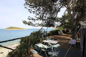 Campingplatz am Meer: Zelt und Campingstühle unter Bäumen, Tisch auf dem Stellplatz, Zaun zum Wasser hin.