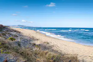 Sandstrand an der Küste mit Wellen im blauen Meer und Dünen mit Gras, dahinter Hügel in der Ferne.