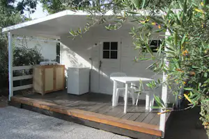 Holzterrasse mit Tisch und weißen Stühlen vor einem weißen Gartenhaus, seitlich ein Grill und umgebende Bäume und Sträucher.