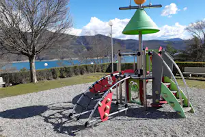 Spielplatz mit Rutsche und Klettergerüst auf einem Campingplatz, im Hintergrund See, Bäume und Berge.