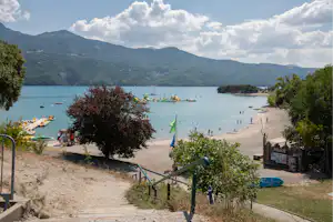 Blick auf einen See mit Sandstrand, Bojen und Bergen im Hintergrund, am Ufer mit Wegen und Bäumen.