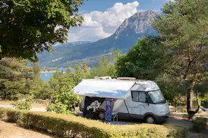 Weißes Wohnmobil mit Markise steht auf einem grasigen Stellplatz, umgeben von Bäumen, mit Bergen im Hintergrund.