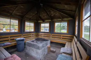 Innenansicht einer Campinghütte mit Holzbänken, Feuerstelle aus Stein und Fenstern zur grünen Umgebung.