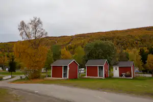 Rote Camping-Unterkünfte mit Schiebetüren am Weg, davor Rasen und Bänke, im Hintergrund bunte Hügel und Bäume.