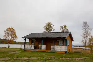 Holzhaus mit Veranda und Geländer am Campingplatz, Rasenfläche und See im Hintergrund, Bäume am Ufer.