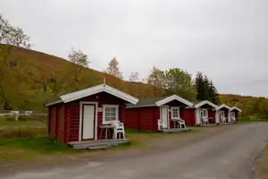 Rote Holz-Häuser mit Veranda und Sitzplätzen am Campingplatz, daneben eine Straße und grüne Hügel im Hintergrund.