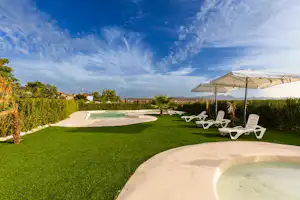 Großer Rasenbereich mit Sonnenliegen unter einem Sonnenschirm, im Vordergrund ein Poolrand, im Hintergrund Hecken und Bäume.