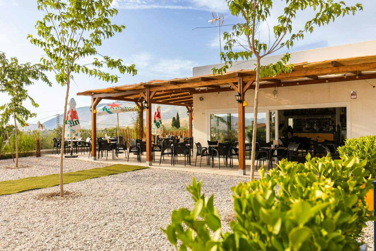 Terrasse mit Holzdach und mehreren Tischen vor einem hellen Gebäude, umgeben von Kiesweg und Bäumen.