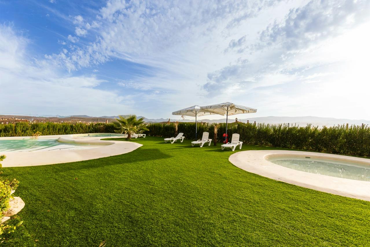 Grüner Rasen mit zwei weißen Liegen und Sonnenschirm, daneben ein Pool mit geschwungenem Rand und Blick auf die Landschaft.