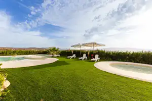 Grüner Rasen mit zwei weißen Liegen und Sonnenschirm, daneben ein Pool mit geschwungenem Rand und Blick auf die Landschaft.