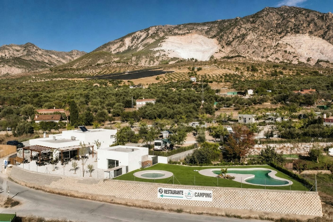 Campinganlage mit Poolbereich und Restaurantgebäude, umgeben von Bäumen und Wohnhäusern, vor einer Berglandschaft im Hintergrund.