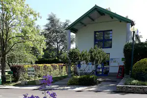 Campinghaus mit grünem Dach, Eingang und Fenstern, davor Weg und Hecken im Garten mit Bäumen.