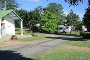 Campingplatz mit Gebäude und grünem Vordach, gepflastertem Weg, Rasen und mehreren Wohnmobilen zwischen Bäumen.