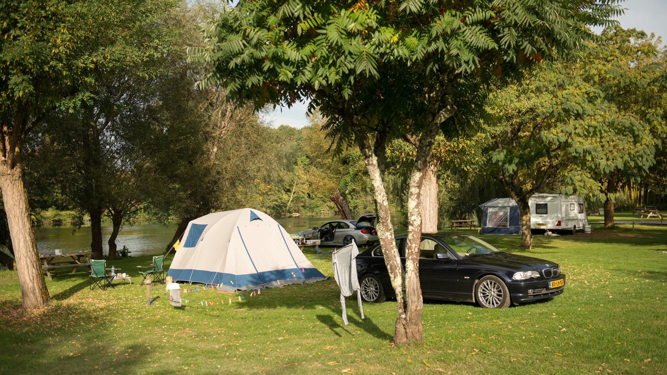 Zelt auf einem Campingplatz mit geparktem Auto und weiteren Wohnwagen im Hintergrund, umgeben von Bäumen am Seeufer.