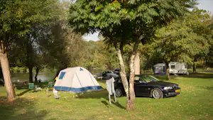 Zelt auf einem Campingplatz mit geparktem Auto und weiteren Wohnwagen im Hintergrund, umgeben von Bäumen am Seeufer.