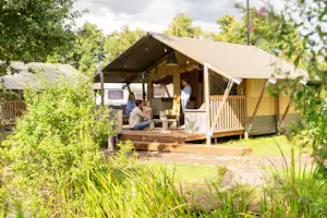 Glamping-Zelt auf Holzterrasse mit Sitzgruppe und Tisch, umgeben von Büschen und Bäumen im Campingbereich.