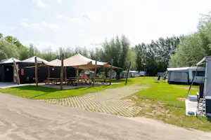 Campingplatz mit Zelt und Sonnensegeln, Holzbank und Tisch unter Bäumen, Stellplätze mit Wohnwagen und Wohnmobilen.