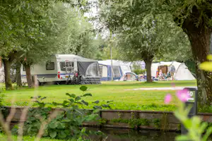 Wohnwagen und Zelte auf einem Campingplatz zwischen Bäumen, mit Fahrrad am Wohnwagen und Campingstühlen am Zelt.